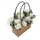 A bouquet of white roses in a brown paper bag with black handles.