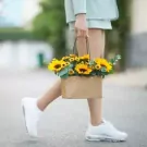 Person in white sneakers carrying a brown gift bag of sunflowers and yellow flowers.