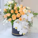 Peach roses, white orchids, and greenery in a dark round box.