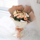 Bouquet of peach roses, white flowers and greenery wrapped in brown and patterned paper with a pink ribbon.