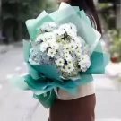 A bouquet of white chrysanthemums and blue hydrangeas wrapped in teal paper.