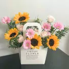 A white floral arrangement with sunflowers, pink gerbera daisies, and light pink roses in a tote bag.