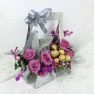 A gift box with purple roses, Ferrero chocolates, and orchids, tied with a silver ribbon.