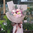Bouquet of pink and purple carnations, roses, and eustoma flowers with greenery, wrapped in beige paper and tied with a brown ribbon.