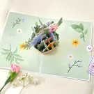 3D pop-up card with a basket of colorful flowers on a light green background with scattered dots and floral illustrations.