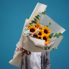 Bouquet of sunflowers and eucalyptus, held by a person in a white shirt and grey cardigan.