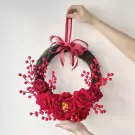 Red rose and berry wreath with a red ribbon bow, held up against a white wall.