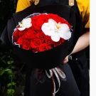 A bouquet of red roses with two white orchids, wrapped in black paper and tied with a white ribbon.
