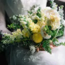 A bridal bouquet of white roses, yellow snapdragons, and greenery held by a bride.