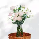 Pink roses and eucalyptus in a glass vase on a woven tray.