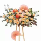 Floral arrangement with orange chrysanthemums, yellow roses, and pink accents on a wooden stand, adorned with paper fans.