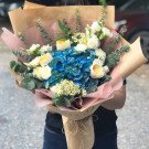 Bouquet of blue hydrangeas, white roses, and small white flowers wrapped in brown paper.