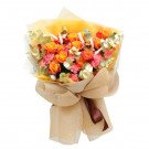 A bouquet of orange roses, pink carnations, and gold accents wrapped in beige paper with a brown ribbon.