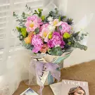 A bouquet of pink peonies, tulips, and lisianthus with eucalyptus and thistles, wrapped in newspaper and sheer fabric, tied with a purple ribbon.