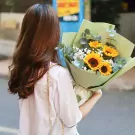 Woman with long brown hair holding a bouquet of sunflowers, baby's breath, and eucalyptus in light green paper.