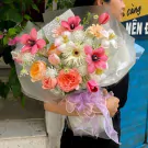 A person holds a colorful bouquet of tulips, roses, and other flowers, tied with a lavender ribbon.