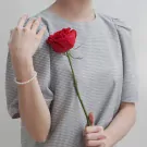Woman in grey gingham dress wearing a pearl bracelet holds a red rose.