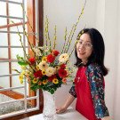 Woman in red shirt with floral sleeves smiles next to a vase of red roses, yellow flowers, and pussy willow branches.