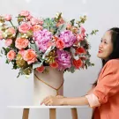 A woman smiles at a large pink and orange floral arrangement in a white box.
