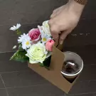 Hand holding a brown cardboard carrier with a bouquet of artificial roses, daisies, and tulips, and a cup of chocolate drink.