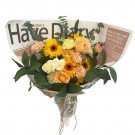 Bouquet of yellow gerbera daisies, orange roses, pink carnations, and greenery wrapped in newspaper.