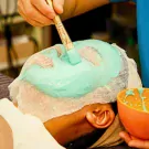 Person receiving a teal facial mask applied with a brush.
