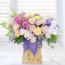 A wooden box filled with pink, purple, and yellow roses, white orchids, and green foliage.