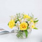 A bouquet of yellow roses and white alstroemeria in a clear glass vase on a white surface.