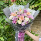 Violet and pink roses, white roses, gerbera daisies, and carnations in a bouquet with grey and white wrapping.