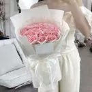 A woman in a light pink dress holds a glamorous pink rose bouquet wrapped in sheer paper and ribbon.