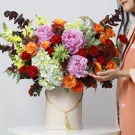 A lush bouquet of pink peonies, orange roses, red carnations, and white hydrangeas in a box.
