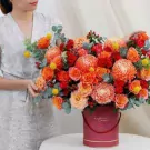 A woman holds a round, maroon hat box overflowing with bright orange roses, chrysanthemums, and yellow billy balls.