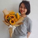 Smiling woman holding a bouquet of vibrant sunflowers wrapped in brown paper with a white ribbon.
