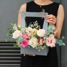 A person holds a pastel flower arrangement in a gray box, featuring roses, lisianthus, and baby's breath.