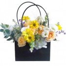 A black gift bag filled with a mixed flower arrangement featuring yellow daisies, peach roses, white roses, and blue hydrangea.