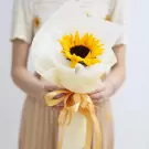 A person holds a bouquet with one sunflower wrapped in white paper and tied with a yellow ribbon.