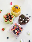 Four beautifully decorated desserts: a mango pudding, a star-shaped green cake with strawberries and blackberries, a heart-shaped chocolate cake with cookies, and a pink flower-shaped cake with mixed berries.