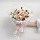 A hand holding a pink and white floral bouquet wrapped in sheer pink paper, tied with a ribbon.