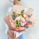 A person in a white shirt and blue jeans holds a bouquet of pink, white, yellow, and orange flowers wrapped in pink paper.