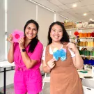 Two women smile, holding pink flower and blue butterfly punch needle crafts.