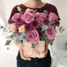 A woman holds a box of pink roses, purple chrysanthemums, and eucalyptus leaves.