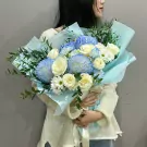 A bouquet of blue and white flowers, including chrysanthemums and roses, wrapped in light blue paper.
