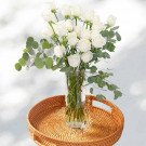 A tall glass vase filled with white roses and eucalyptus sits on a woven tray.