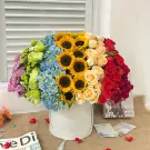 Round flower box with rainbow arrangement of sunflowers, hydrangeas, and roses in red, cream, and purple.