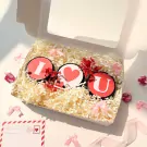 Gift set with three round candles spelling "I Heart U" nestled in shredded paper, surrounded by pink bows and dried flowers.