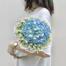 A woman in a white t-shirt holds a bouquet of blue hydrangeas and white daisies trimmed with pearls.