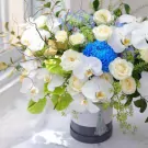 White orchids, roses, and blue chrysanthemum in a grey and white floral box.