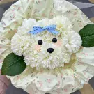 White flower arrangement shaped like a puppy's face with a blue bow, on floral wrapping paper.