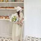 A woman in a cream dress and white hat holds a bouquet of white and yellow flowers.