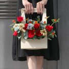A cream tote bag filled with red roses, grey roses, and daisies is held by a person in a black dress.